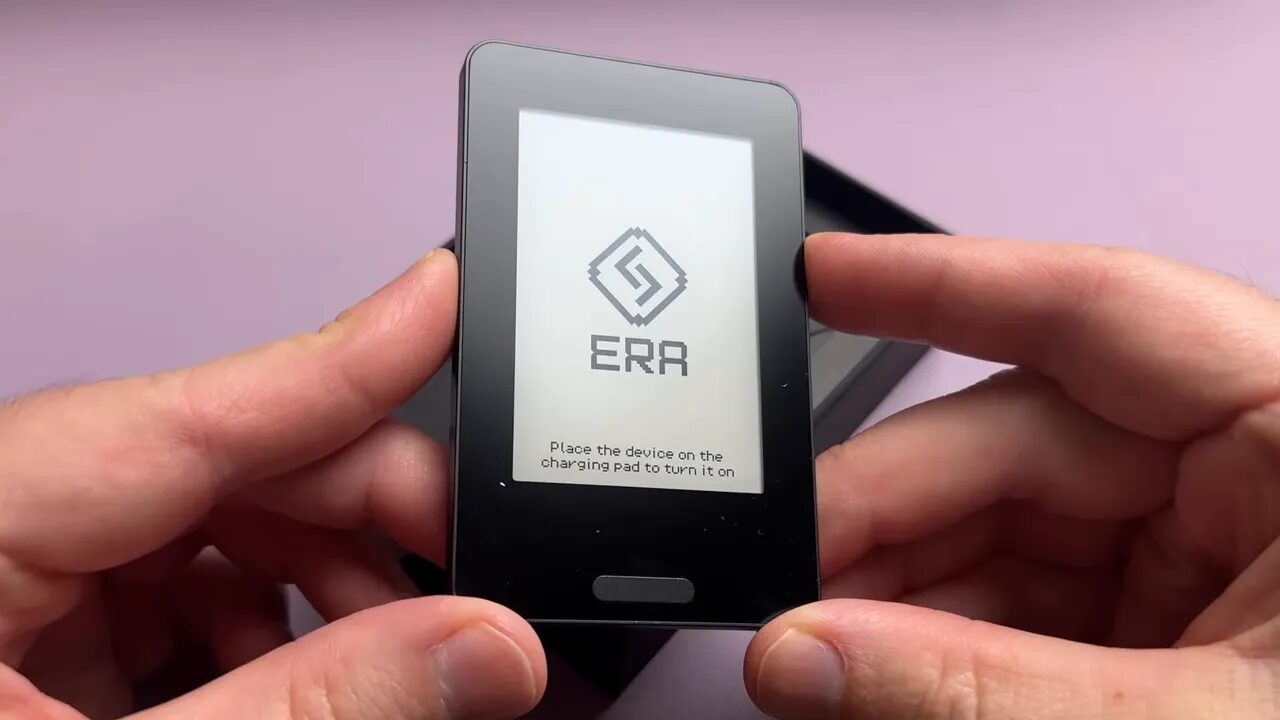 Front view of the Era hardware wallet showing the Era logo and the instruction 'Place the device on the charging pad to turn it on', held between two hands.