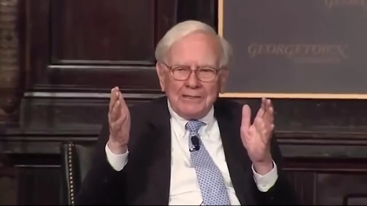 Seated speaker in suit and tie facing camera with hands raised, clear mid-shot
