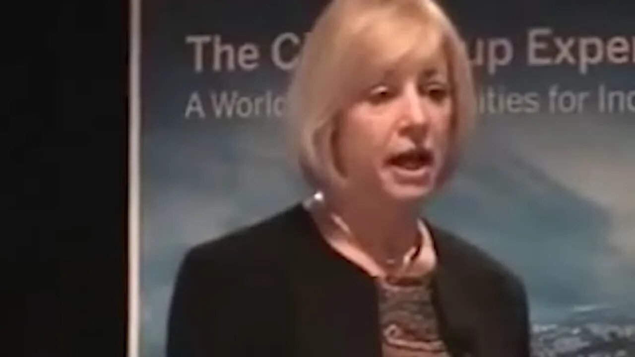 Frontal mid-shot of a woman speaker addressing an audience, conference banner behind