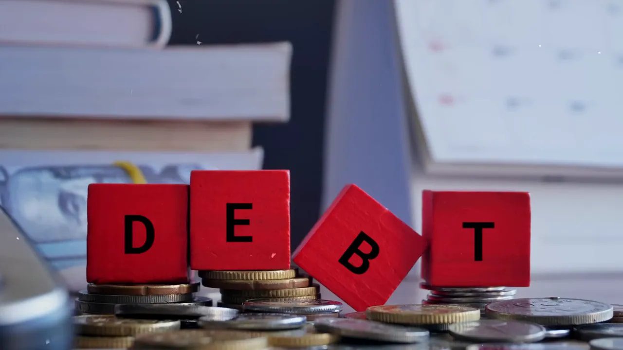 red wooden blocks spelling 'DEBT' placed on stacks of coins, with books and a calendar blurred in the background